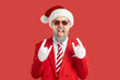 © Studio Romantic - Let's party. Bearded man in red suit, striped tie, Christmas Santa cap, white gloves and sunglasses standing on red background doing rock hand sign and sticking tongue out with excited face expression