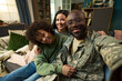 © pressmaster - Portrait of Black middle aged man in military uniform sitting on sofa with smiling Caucasian woman and curly haired child, family members embracing and looking at camera together