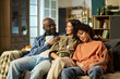© pressmaster - Black middle aged man in military uniform sitting on couch with smiling Latina young adult woman and sleeping Black teenager, family members embracing each other at home