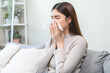 © KMPZZZ - Sick, coronavirus or covid, tired asian young woman have a fever, flu and hand in use tissues paper sneezing nose, runny while sitting on sofa, couch at home. Health care with disease, pandemic virus