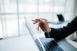 © amnaj - Businesswoman holding a pen and pointing at a laptop screen while engaged in work at a bright, modern office environment