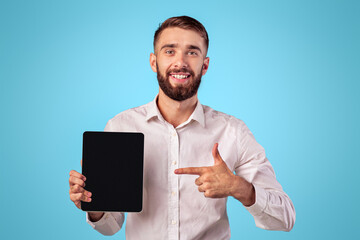Young businessman pointing at tablet with empty screen on blue studio background, mockup for application or website design. Cheerful millennial guy showing touch pad with space for your ad