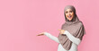 © Prostock-studio - Beautiful muslim woman in hijab demonstrate something on her empty palm and pointing at it, standing over pink background, panorama with copy space