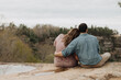 © Jordan - Man and woman sitting side by side on a rocky cliff edge overlooking a forest valley with arm around in a serene romantic outdoor moment
