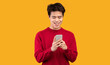 © Prostock-studio - People And Technology Concept. Portrait of happy smiling young asian man using smartphone standing isolated over orange studio background, copy space. Guy chatting online, browsing social media