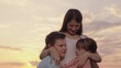 © Victoriia - Family, young mother hugging children, boy girl in park against sky. Happy children daughter son hugging mommy in summer park. Carefree childhood, joyful hugs of mother child. Happy family in nature