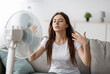 © Prostock-studio - Sad young european woman suffers from unbearably too hot weather, catches cold air from fan in living room interior. Home without air conditioning, lady tired from summer heat, waves arms to cool down