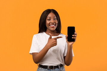 Smiling young black lady showing modern mobile phone, pointing at empty screen, sharing her positive emotions after using nice mobile application, orange studio background, copy space