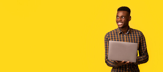 Remote job for students. Portrait of happy young black guy with laptop computer in hands looking aside at copy space, standing on yellow background, millennial African man enjoying freelance work