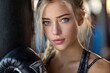 © Kien - A beautiful blonde woman with blue eyes, wearing black boxing gloves and standing next to a punching bag in the gym, looks directly at the camera
