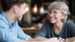 © Curioso.Photography - A smiling senior woman with glasses joyfully engages in conversation with a younger man, sharing a warm intergenerational moment.