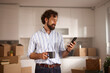 © Stockphotodirectors - A man stands in his new home, smiling as he looks at his phone while holding a coffee cup. Moving boxes are stacked around him, suggesting he is in the process of settling in.