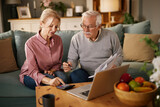 Two senior adults, a man and a woman, sit on a couch at home. They review paperwork and use a calculator with a laptop to discuss their life insurance policy.