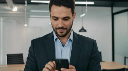 A smiling businessman stands in an office, looking at his phone