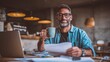© Attasit - Man enjoying coffee while working, looking at camera, laptop in front, paper documents on desk.