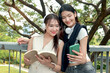 © Stella - Happy two beautiful Asian women use mobile phone to take a selfie photo together outdoor in park. Smiling close friends hold a book, spend time outside together in green garden. People in nature