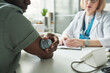 © pressmaster - Black middle aged man checking blood glucose with medical device while sitting across from Caucasian middle aged female doctor in medical office during diabetes consultation