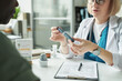 © pressmaster - Middle aged Caucasian female doctor demonstrating blood glucose meter to middle aged Black man during diabetes consultation in medical office, medical form and diabetes chart visible on desk