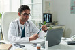 © pressmaster - Middle aged Caucasian male doctor sitting at desk examining insulin pen while consulting diabetic patient in medical office, laptop and medication bottle visible on table