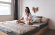 © BullRun - Smiling young woman meditates in lotus pose on bed, radiating warmth, mindfulness, and modern spiritual wellness in serene minimalist home interior.