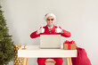 © Studio Romantic - Funny modern Santa working in his office. Happy man in red suit, Xmas cap and cool sunglasses gives thumbs up while sitting at desk with laptop computer next to Christmas tree on light wall background