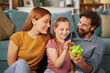 © Stockphotodirectors - A family sits together on a couch, sharing smiles and laughter as they help a young girl count coins in a colorful piggy bank during a cozy evening at home.