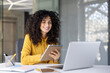 © Liubomir - A smiling woman with curly hair takes notes while working on a laptop in a modern office setting.