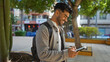 © Krakenimages.com - Young man using a tablet while smiling outdoors in a vibrant urban park setting, showcasing modern technology and casual lifestyle in a lively city environment.
