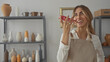 © Krakenimages.com - Blonde woman holds smartphone to mouth with bare arms in pottery workshop; joy laughter creativity enthusiasm.