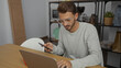 © Krakenimages.com - Young man in office working on laptop and phone focused on woodworking shelves behind relaxed setting professional attire.