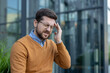 © Tetiana - Close-up photo of a young businessman sitting tiredly outside an office building, hiding from the pain and holding his head with his hand