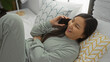 © Krakenimages.com - Woman talking on phone in cozy bedroom shows a young asian female enjoying a conversation at home, relaxed on a bed with patterned pillows in a modern interior setting.