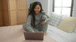 © Krakenimages.com - Woman sitting on bed using laptop and phone, showcasing a relaxed indoor atmosphere with light filtering through the window, focusing on work from home lifestyle.
