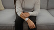 © Krakenimages.com - Woman sitting on a gray sofa in a living room scratching her arm wearing a white sweater and watch, suggesting an itchy sensation in a cozy home setting.