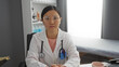 © Krakenimages.com - Woman doctor sitting in hospital clinic wearing a white coat with glasses showcasing a professional asian medical workplace with stethoscope and clipboard.