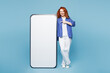 © ViDi Studio - Full body young ginger woman she wear shirt t-shirt casual clothes point finger on big huge blank screen mobile cell phone smartphone with workspace area isolated on plain light blue cyan background.