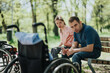 © qunica.com - A serene image of a couple leisurely sitting together on a park bench with two wheelchairs nearby, surrounded by natural greenery and sunlight.