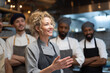 © Katie R - female head chef gesturing while briefing diverse kitchen staff, lively team atmosphere