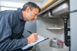 © Mohdfaridzazhar - Inspector examining plumbing under the kitchen sink with flashlight and checklist,