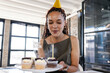 © Wavebreak Media - African American woman wearing party hat sitting at desk holding frosted cupcakes with lit candle