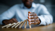 © Sergio IQstock - a professional businessman stopping a falling domino chain with his hand, close-up to medium-close composition, foreground focused on a realistic human hand reaching from the right side of the frame