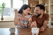 © Migma_Agency - Happy couple saving money inserting coins in piggy bank at home
