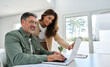 © insta_photos - Happy mature older family couple using laptop technology at home. Smiling middle aged senior man working on computer sitting at table with wife standing nearby in living room talking buying online.