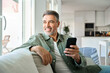 © insta_photos - Relaxed happy mature middle aged man holding cell mobile phone using smartphone sitting at home on sofa, relaxing scrolling social media, checking financial app, buying online, looking away at window.