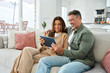 © insta_photos - Middle aged man and woman hugging sitting on sofa at home together relaxing on couch using digital tablet computer. Happy mature old couple buying online browsing internet in modern house living room.