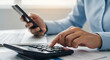 © Stock Studio - Person using a calculator while holding a phone with a laptop visible in the background on a desk