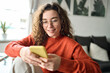 © insta_photos - Happy girl checking cell phone online education apps, texting, browsing internet for shopping sitting at home. Young smiling woman wearing glasses holding smartphone using cellphone, looking at mobile