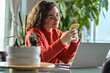 © insta_photos - Happy young lady using smartphone social media apps at home. Smiling woman holding cellphone modern ai applications, looking at mobile, checking cell phone, texting chatting sitting at table at home.
