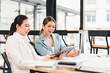 © kenchiro168 - Two businesswomen discussing work while sitting at table with documents and tablet in modern office, showing focus and collaboration in bright environment
