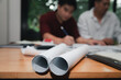 © skarie - Rolled blueprints on a wooden table with blurred architects in the background discussing a project. Concept of architecture, planning, and teamwork in a modern office.
